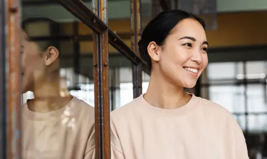 A woman smiling warmly, with her reflection visible in a mirror beside her, wearing a beige sweater.