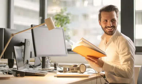 A man in an office sitting down and smiling while holding a book