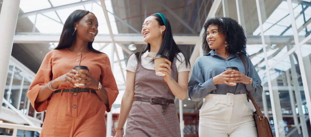 Three women smiling and holding coffee cups, walking together in a modern glass building.